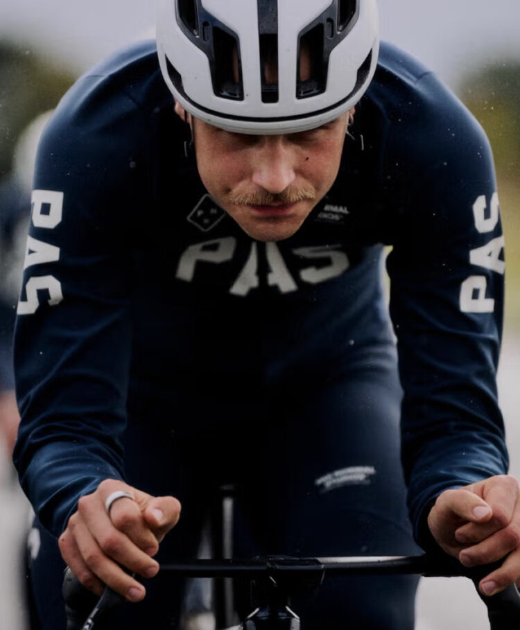 A cyclist wearing a white helmet and dark Pas Normal Studios PAS AW25 Collection cycling gear labeled "PAS" rides in a racing position, gripping the handlebars and looking ahead.