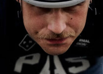 A cyclist wearing a white helmet and dark Pas Normal Studios PAS AW25 Collection cycling gear labeled "PAS" rides in a racing position, gripping the handlebars and looking ahead.