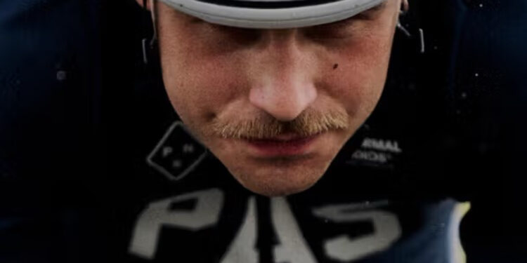 A cyclist wearing a white helmet and dark Pas Normal Studios PAS AW25 Collection cycling gear labeled "PAS" rides in a racing position, gripping the handlebars and looking ahead.