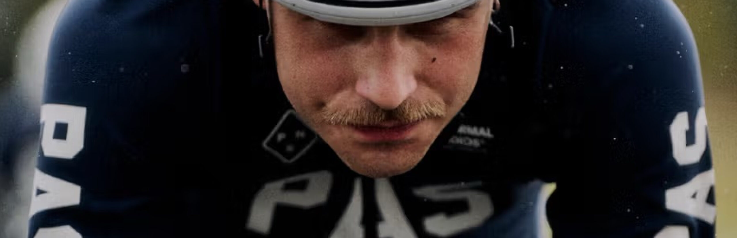 A cyclist wearing a white helmet and dark Pas Normal Studios PAS AW25 Collection cycling gear labeled "PAS" rides in a racing position, gripping the handlebars and looking ahead.